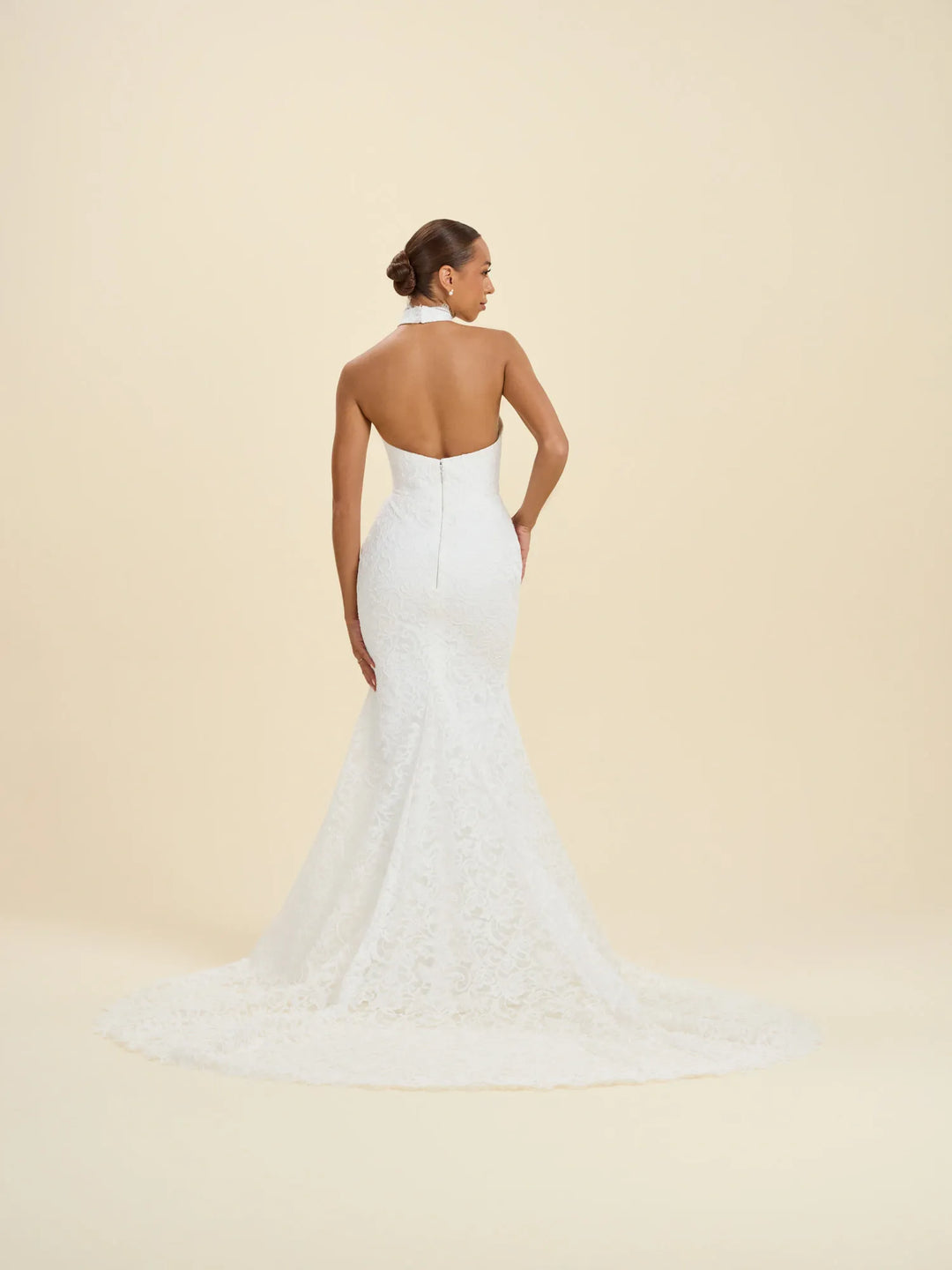 A woman modeling a white lace bridal gown from the back, showing a halter neckline, open back, fitted silhouette, and long train against a warm cream studio background.