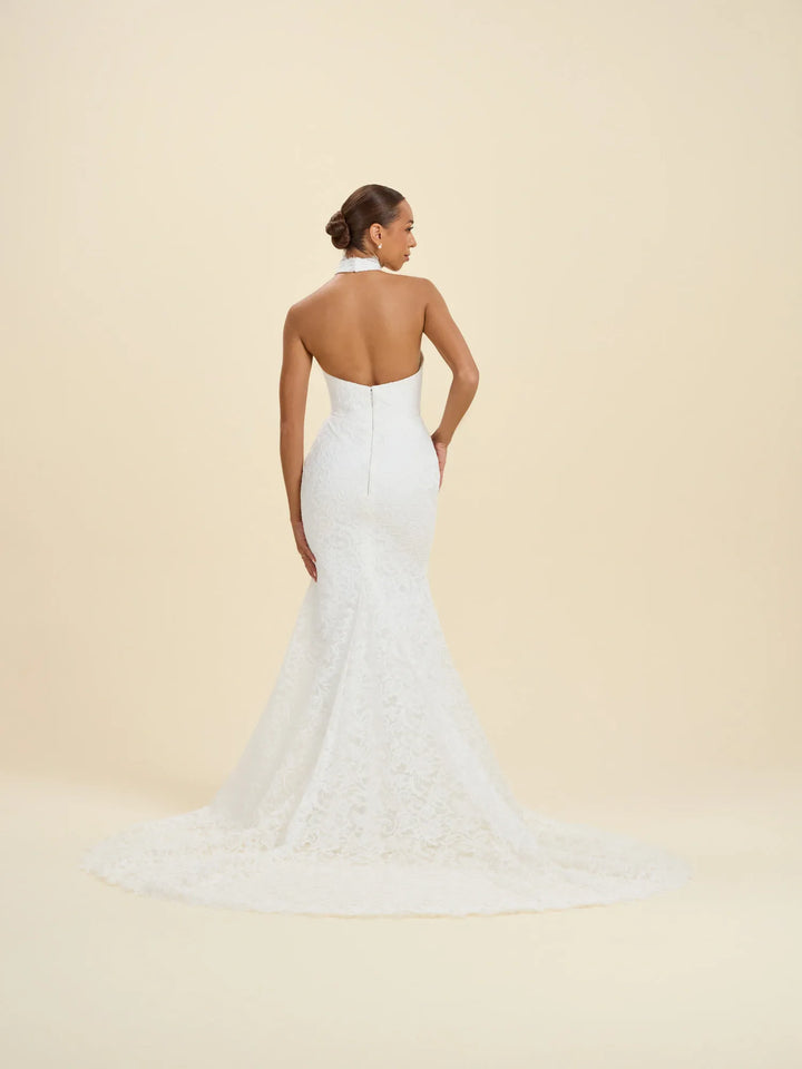 A woman modeling a white lace bridal gown from the back, showing a halter neckline, open back, fitted silhouette, and long train against a warm cream studio background.