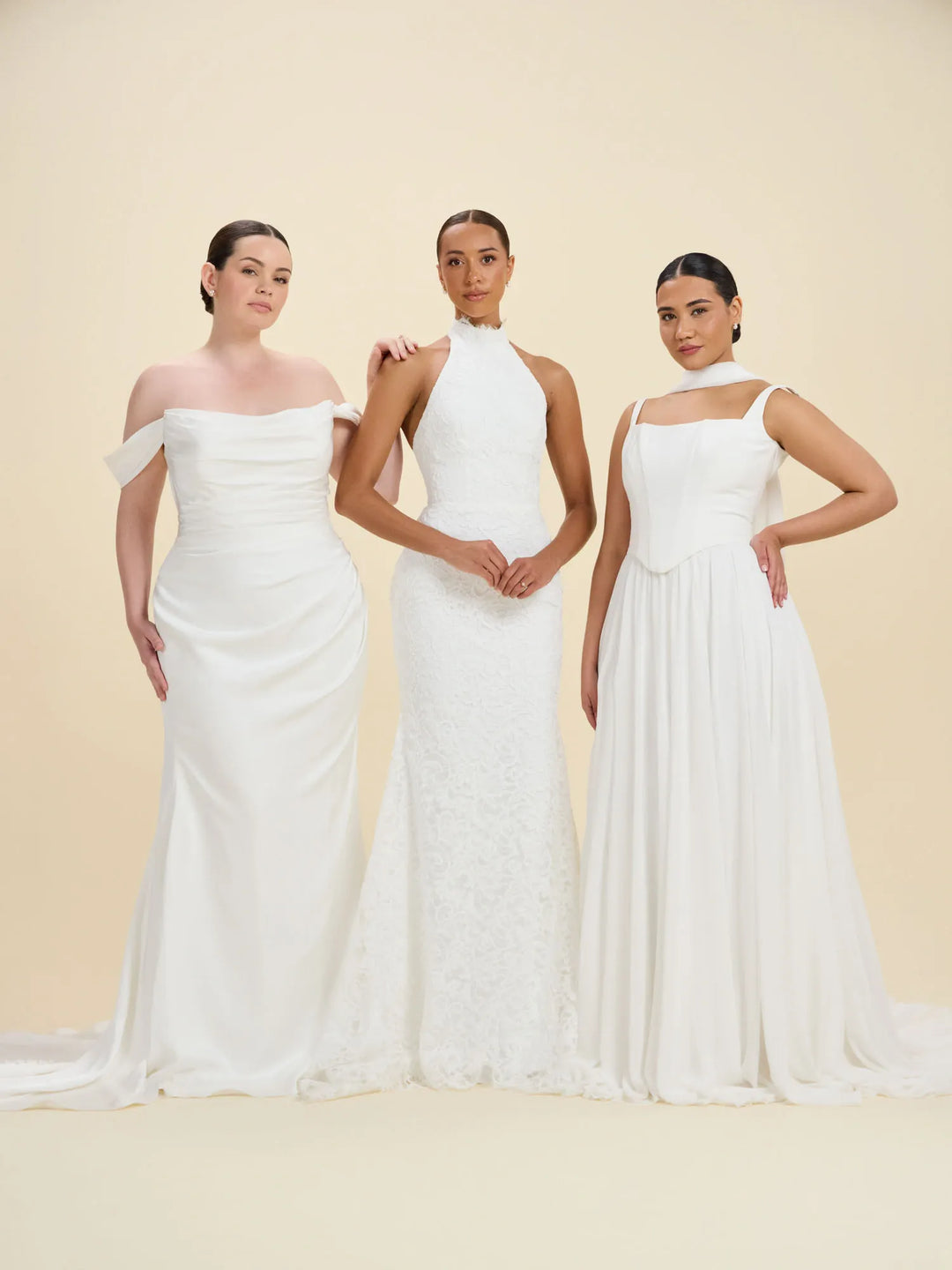 Three women modeling white wedding dresses against a warm cream studio backdrop. The left dress is an off-the-shoulder satin gown with a slight train.