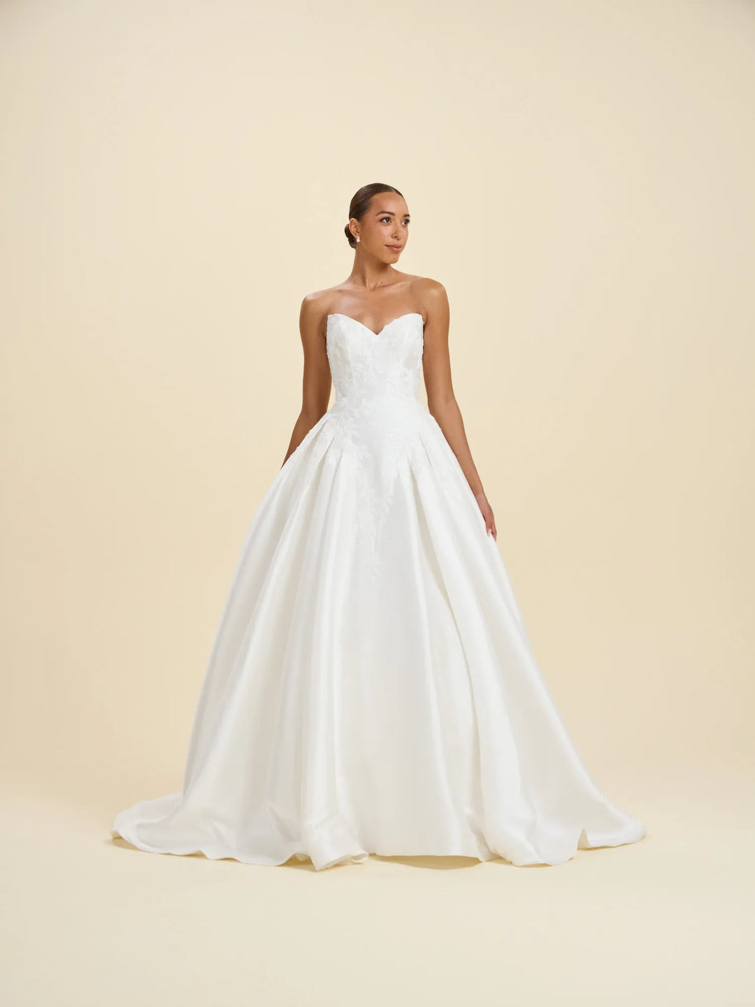 A woman modeling a strapless white wedding gown with a sweetheart neckline, fitted bodice, and full satin A-line skirt against a warm cream studio backdrop.