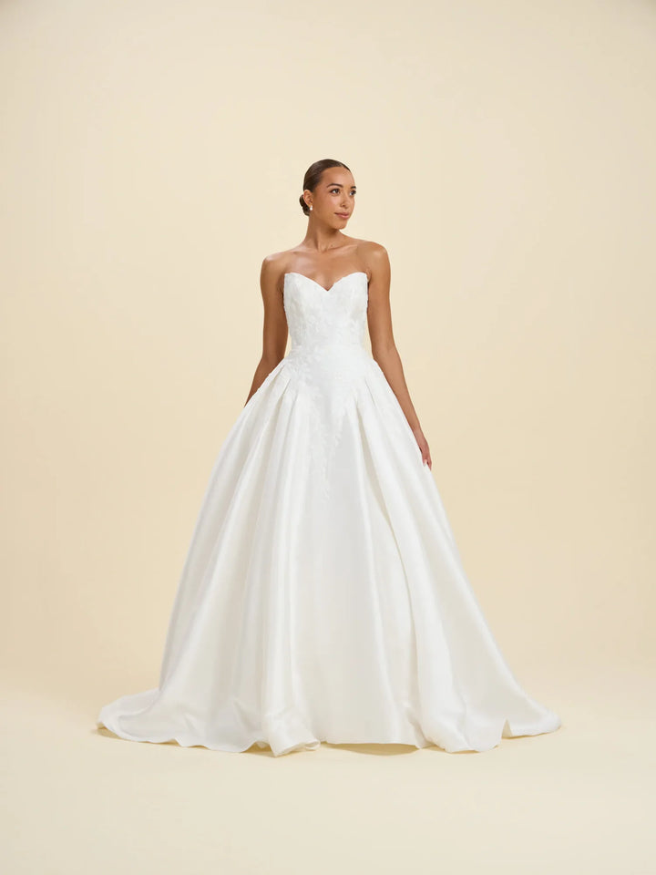 A woman modeling a strapless white wedding gown with a sweetheart neckline, fitted bodice, and full satin A-line skirt against a warm cream studio backdrop.