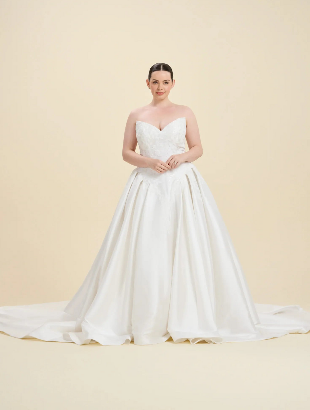 A woman modeling a strapless white wedding gown with a sweetheart neckline, fitted bodice, and full satin train against a warm beige studio background.