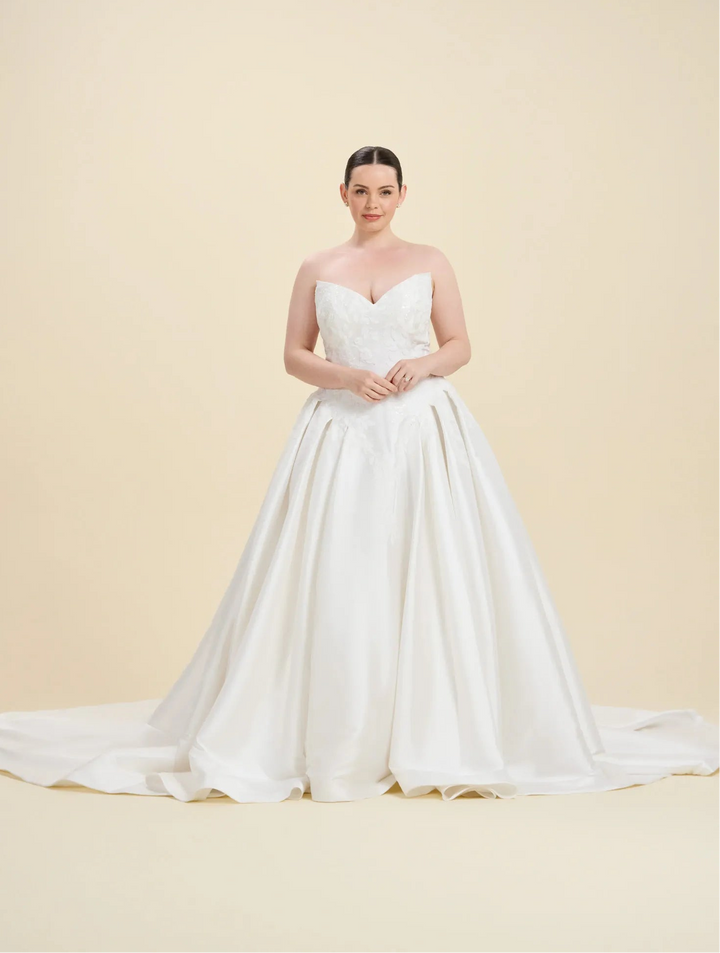 A woman modeling a strapless white wedding gown with a sweetheart neckline, fitted bodice, and full satin train against a warm beige studio background.