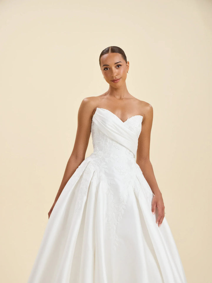 A woman modeling a strapless white wedding gown with a sweetheart neckline, ruched bodice, and full ball skirt against a warm beige studio background.