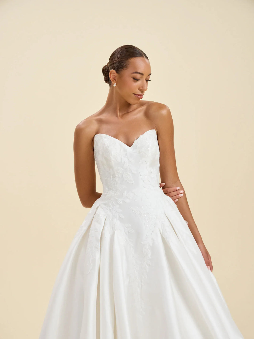 A woman modeling a strapless white wedding gown with a sweetheart neckline, fitted lace bodice, and full satin skirt against a warm beige studio background.