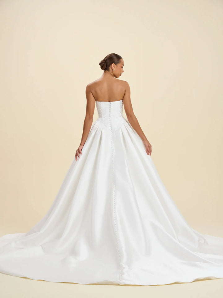 A woman modeling a strapless white wedding gown from the back, showing a structured bodice and a full satin train against a warm beige studio background.