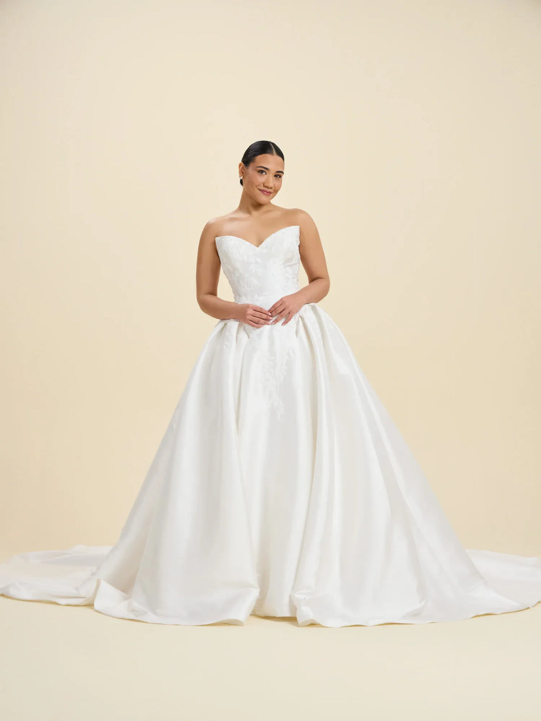 A woman modeling a strapless white wedding gown with a sweetheart neckline, fitted bodice, and full satin train against a warm cream studio backdrop.