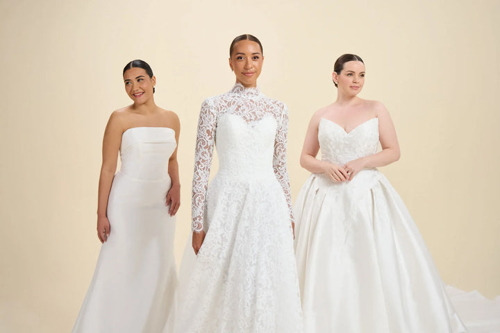 Three women modeling white wedding dresses against a plain beige studio background. The woman in the center wears a long-sleeve lace wedding gown with a high neckline, while the woman on the left wears a strapless white gown and the woman on the right wears a strapless A-line wedding gown.
