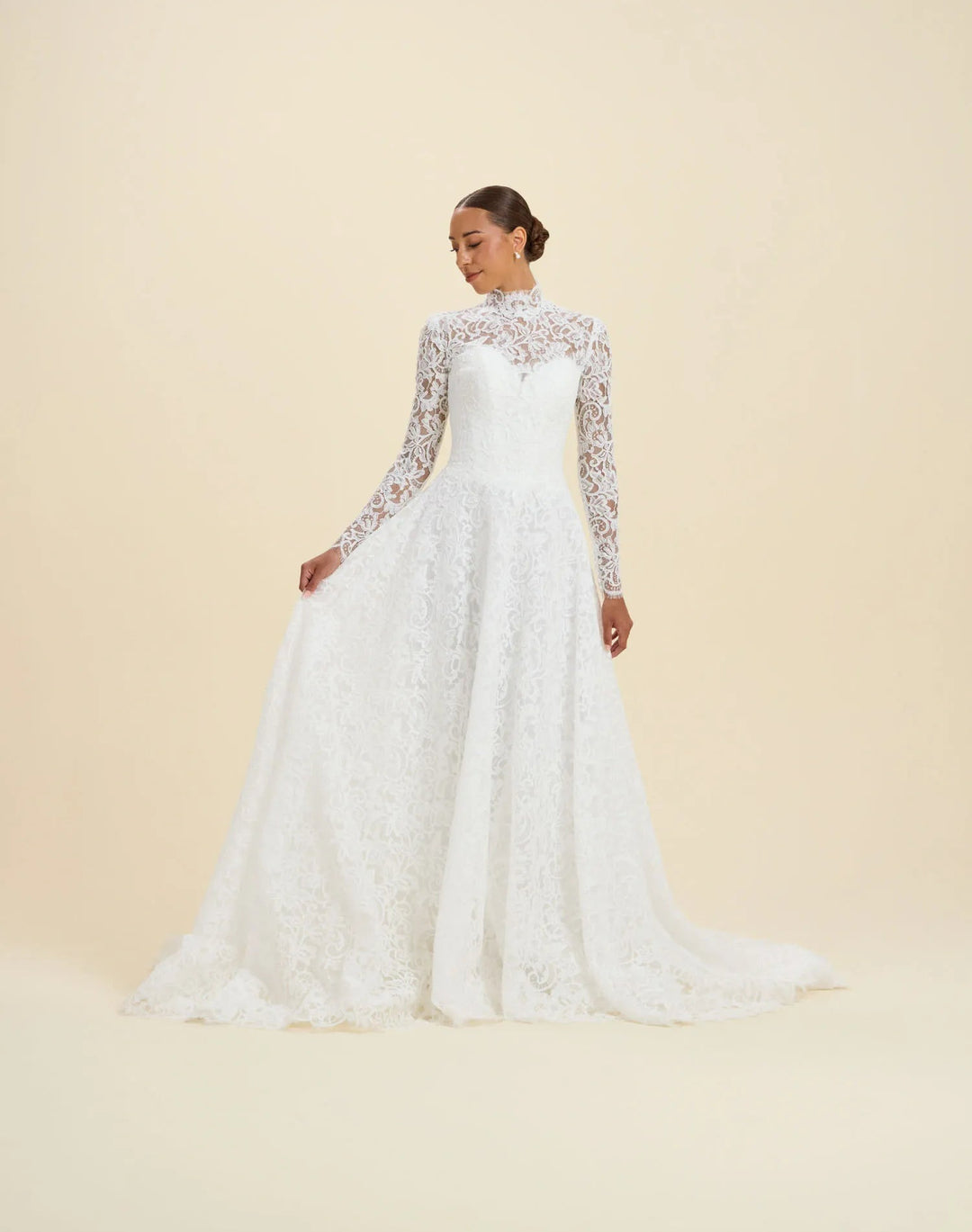 A woman modeling a long-sleeve white lace wedding gown with a fitted bodice and full skirt, photographed against a plain warm beige studio background.