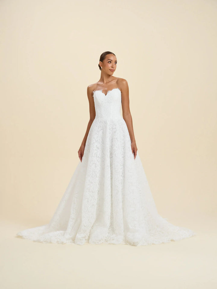 A woman modeling a strapless white lace wedding gown with a sweetheart neckline and a full A-line skirt, photographed against a plain warm beige studio background.