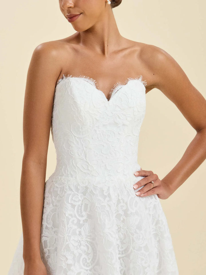 A close-up of a woman wearing a strapless white lace wedding dress with a sweetheart neckline and a flared skirt, posed against a warm beige studio background.
