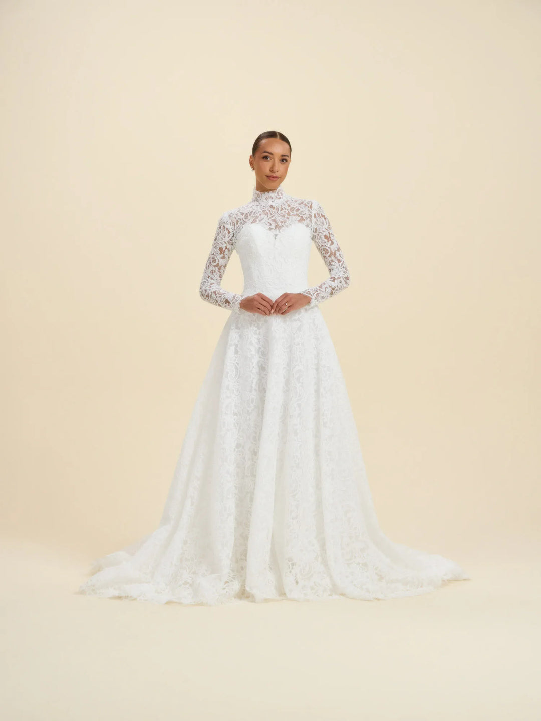 A woman modeling a white lace wedding gown with a high mock neck, long sheer lace sleeves, a fitted bodice, and a full skirt with a slight train, photographed against a plain warm beige studio background.