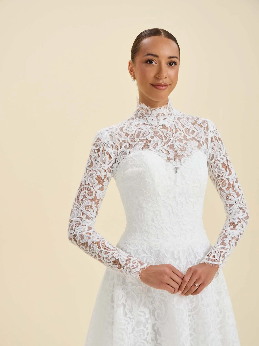 A woman modeling a white lace long-sleeve wedding dress with a high mock neck and fitted bodice against a plain beige studio background.