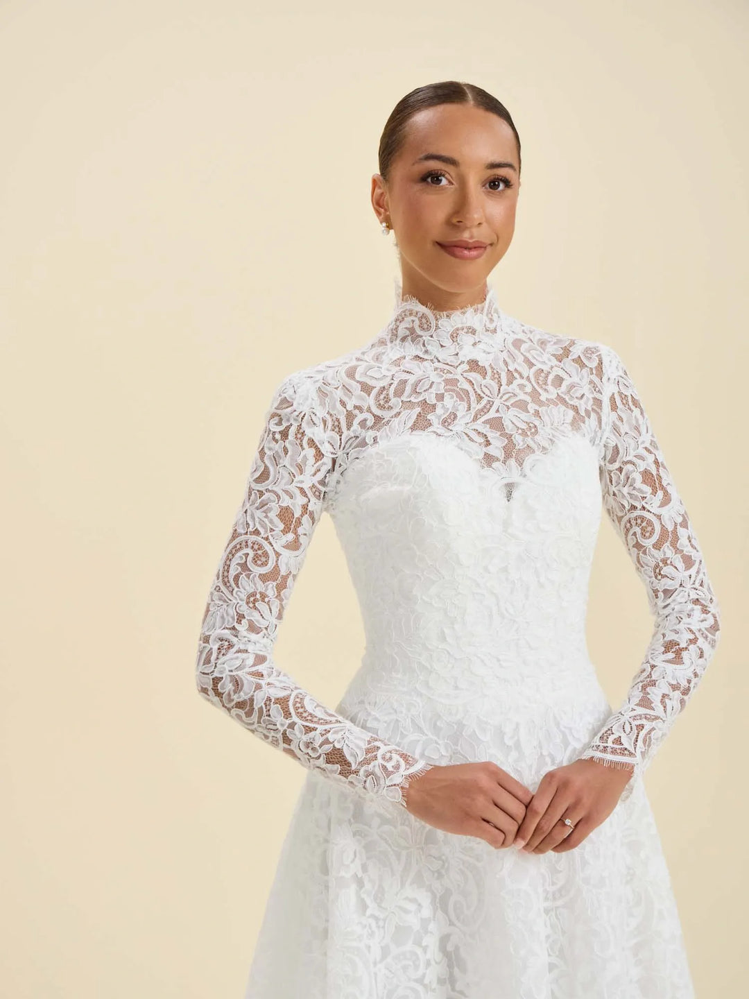 A woman modeling a white lace long-sleeve wedding dress with a high mock neck and fitted bodice against a plain beige studio background.