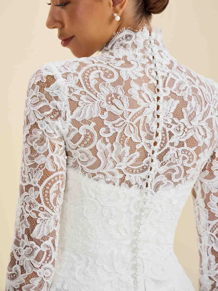 Close-up back view of a woman wearing a white lace bridal dress with long sleeves, floral lace overlay, and a high neckline against a soft beige background.