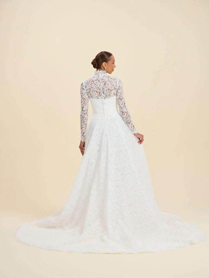 A woman modeling a white lace wedding gown from the back, showing long lace sleeves, a high lace neckline, a fitted waist, and a full lace skirt with a train, against a warm cream studio background.