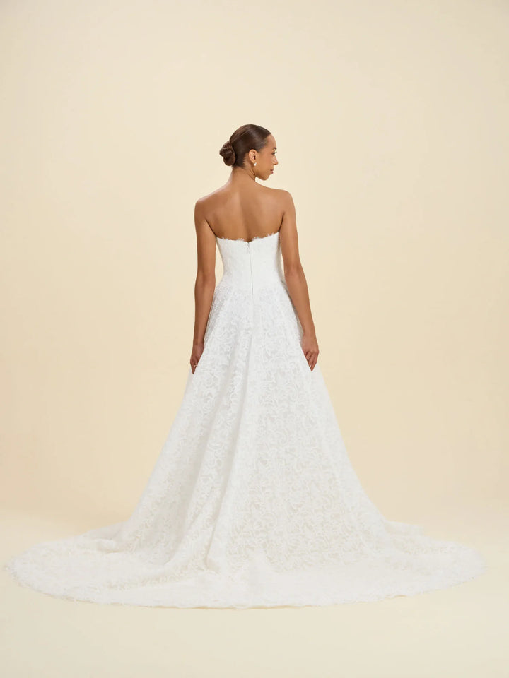 A woman modeling a strapless white lace wedding gown from the back, showing a fitted bodice and full flared skirt with a long train, against a plain warm beige studio background.