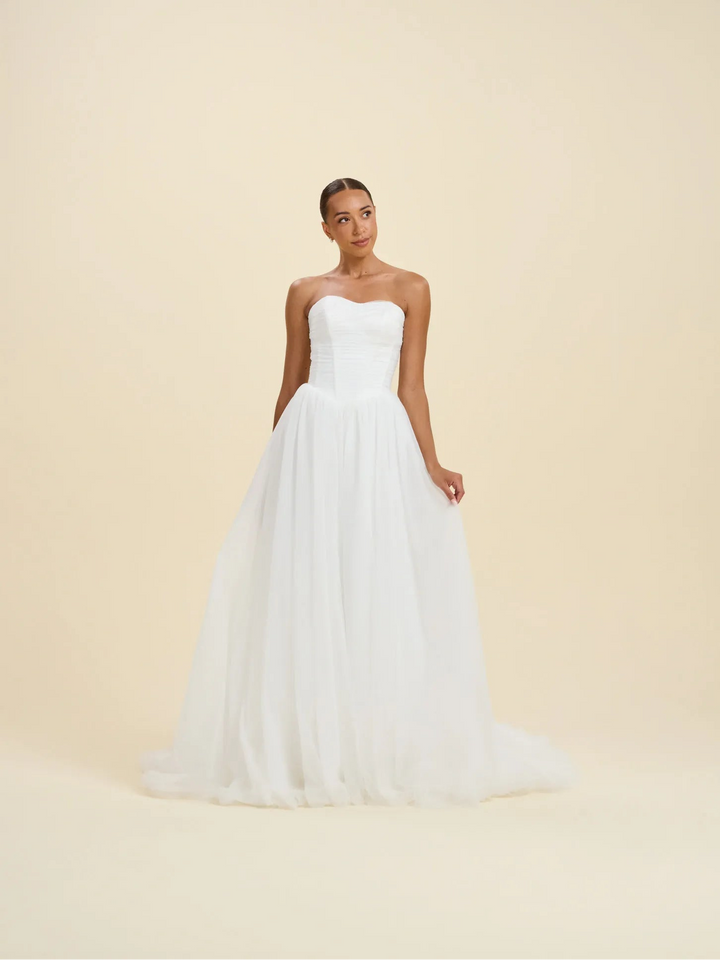 A woman modeling a strapless white wedding gown with a fitted bodice and a voluminous tulle skirt, standing against a plain warm beige studio background.