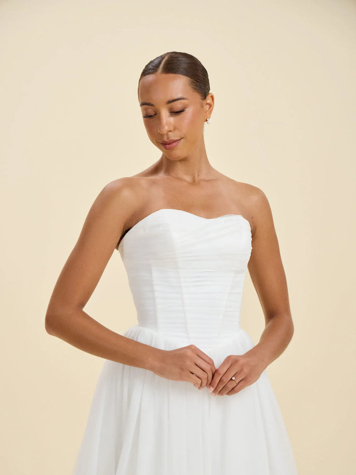A woman wearing a strapless white wedding dress with a structured, ruched bodice and a flowing skirt, standing against a solid beige studio background.