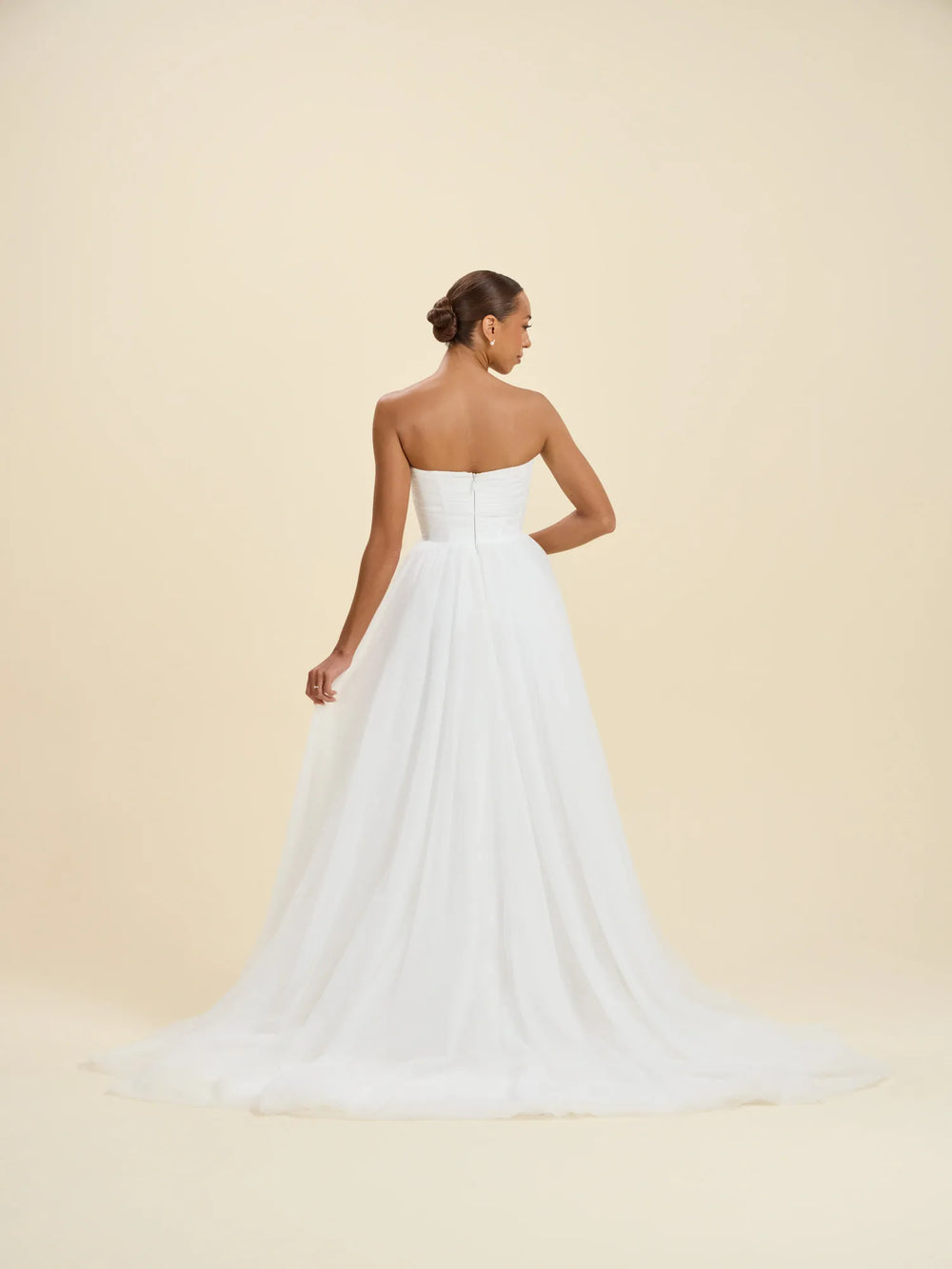 A woman modeling a strapless white wedding gown from the back, showing a structured bodice and a full flowing skirt with a train, against a plain warm beige studio background.