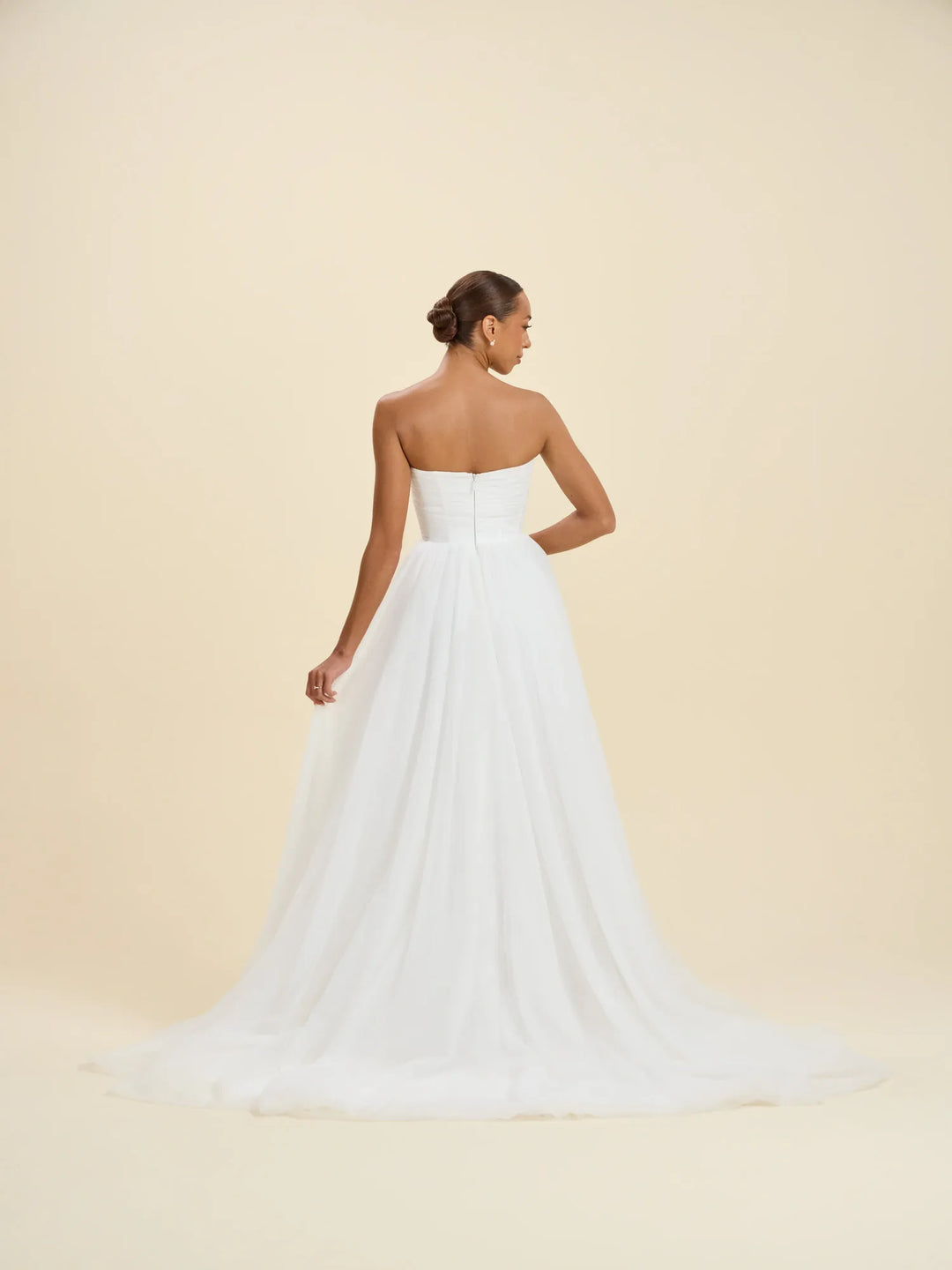 A woman modeling a strapless white wedding gown from the back, showing a structured bodice and a full flowing skirt with a train, against a plain warm beige studio background.