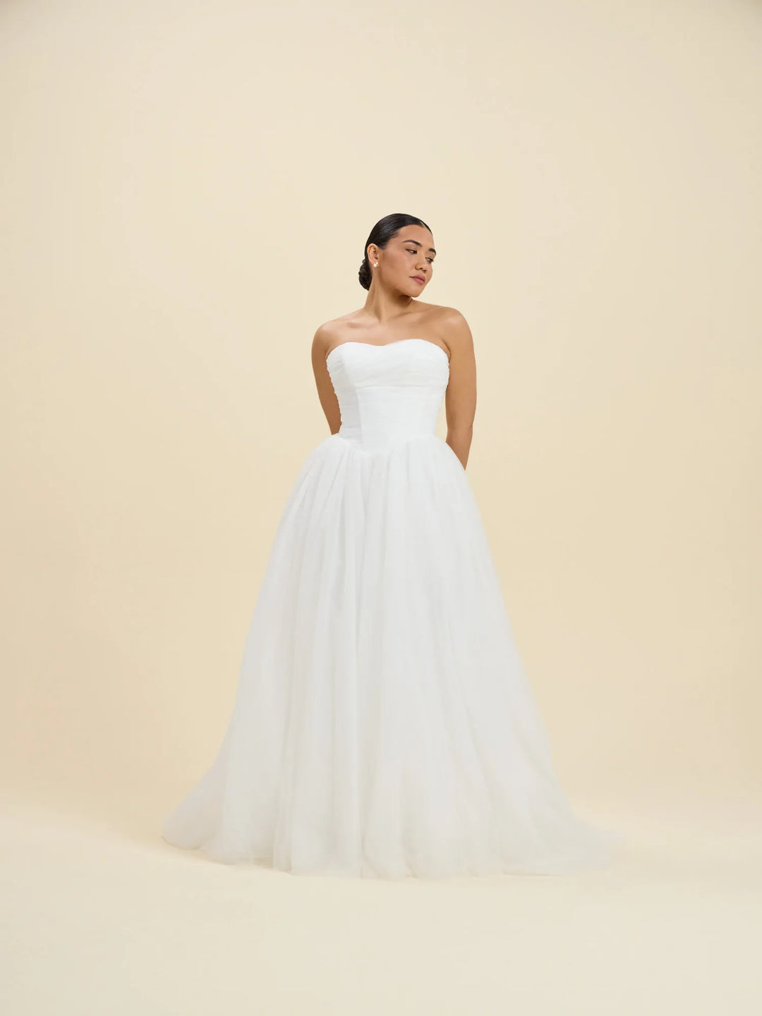 A woman modeling a strapless white wedding gown with a fitted bodice and a voluminous tulle skirt, standing against a plain warm beige studio background.
