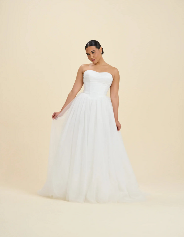 A woman modeling a strapless white wedding gown with a fitted bodice and a full, flowing tulle skirt against a plain warm beige studio background.