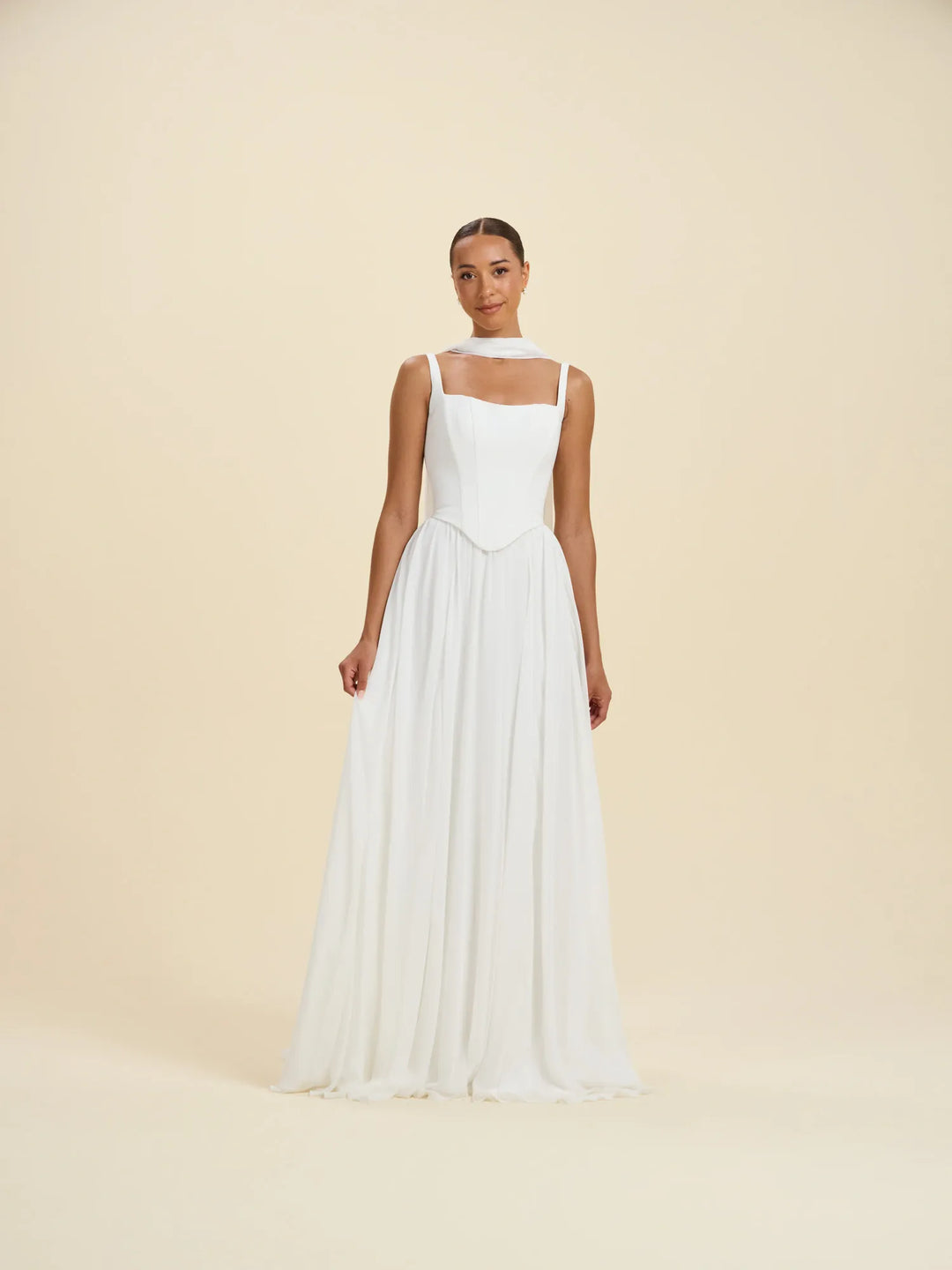 A woman modeling a sleeveless white formal gown with a structured bodice, sheer neckline, and a flowing floor-length skirt, photographed against a plain warm cream studio background.