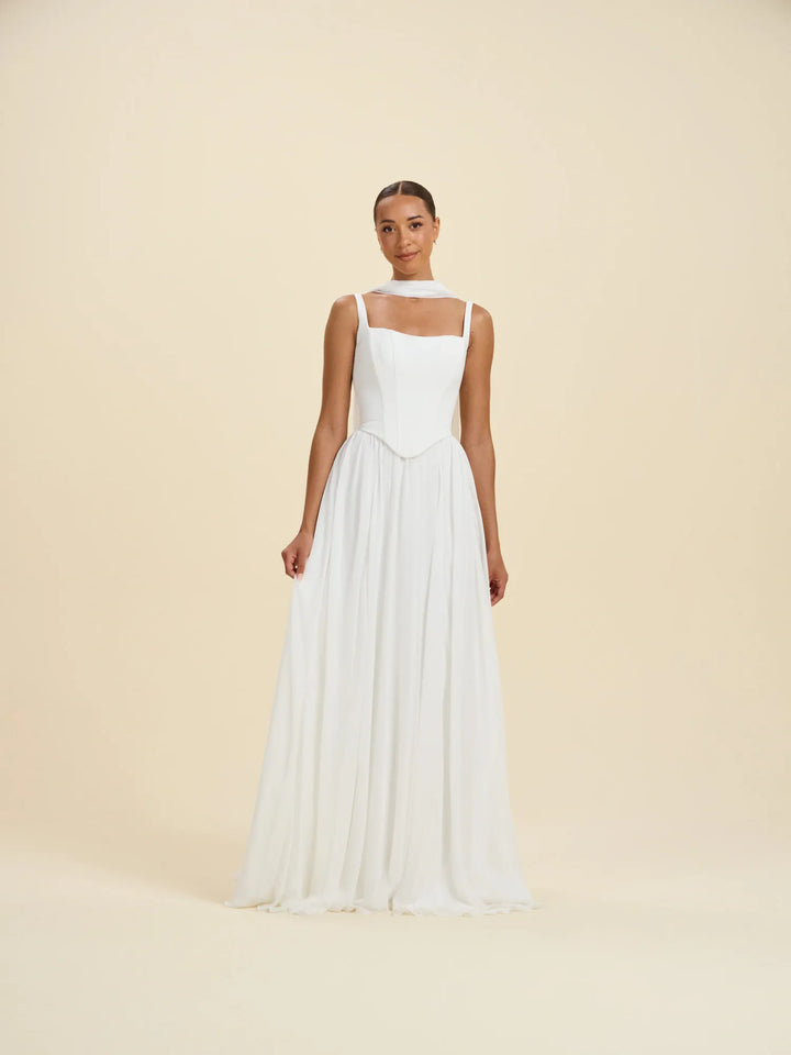A woman modeling a sleeveless white formal gown with a structured bodice, sheer neckline, and a flowing floor-length skirt, photographed against a plain warm cream studio background.