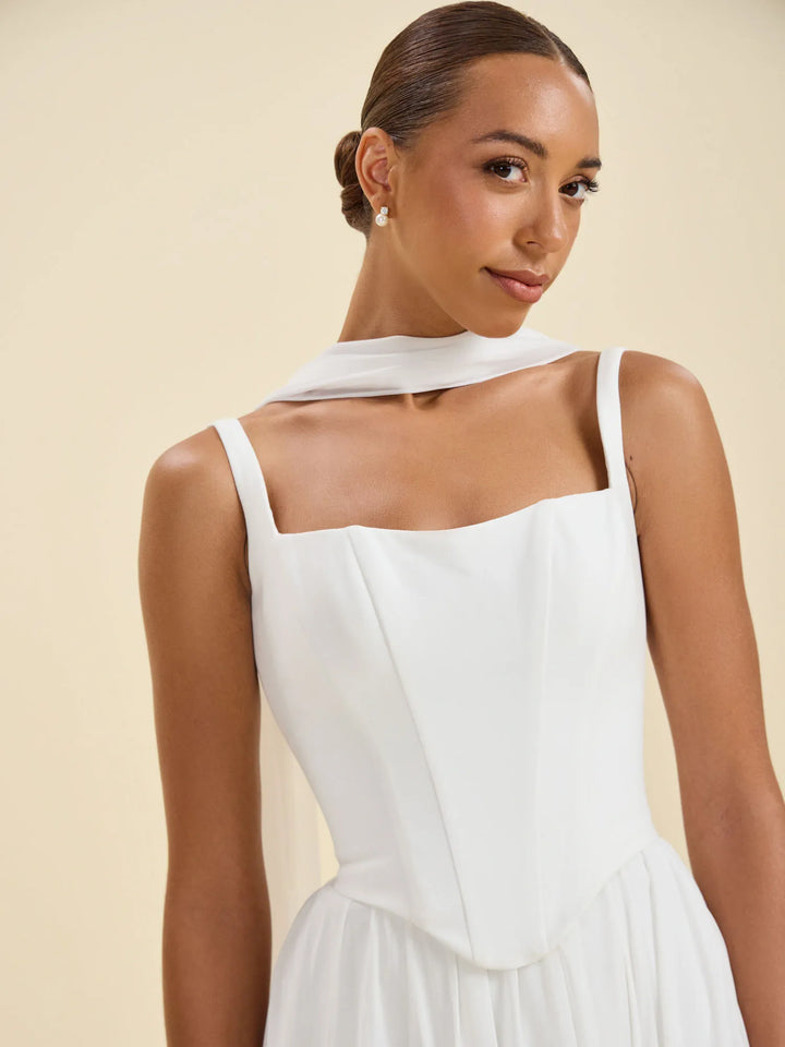 A woman wearing a white sleeveless dress with a draped high neckline and structured bodice, photographed against a warm beige studio background.
