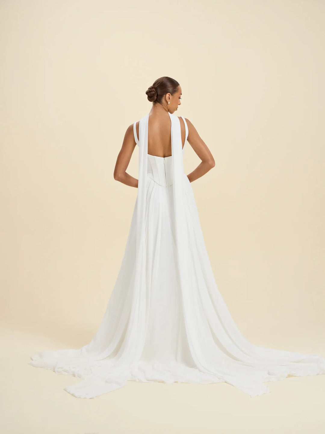 A woman modeling a white sleeveless gown from the back, showing a deep open back and long flowing train, against a warm beige studio background.