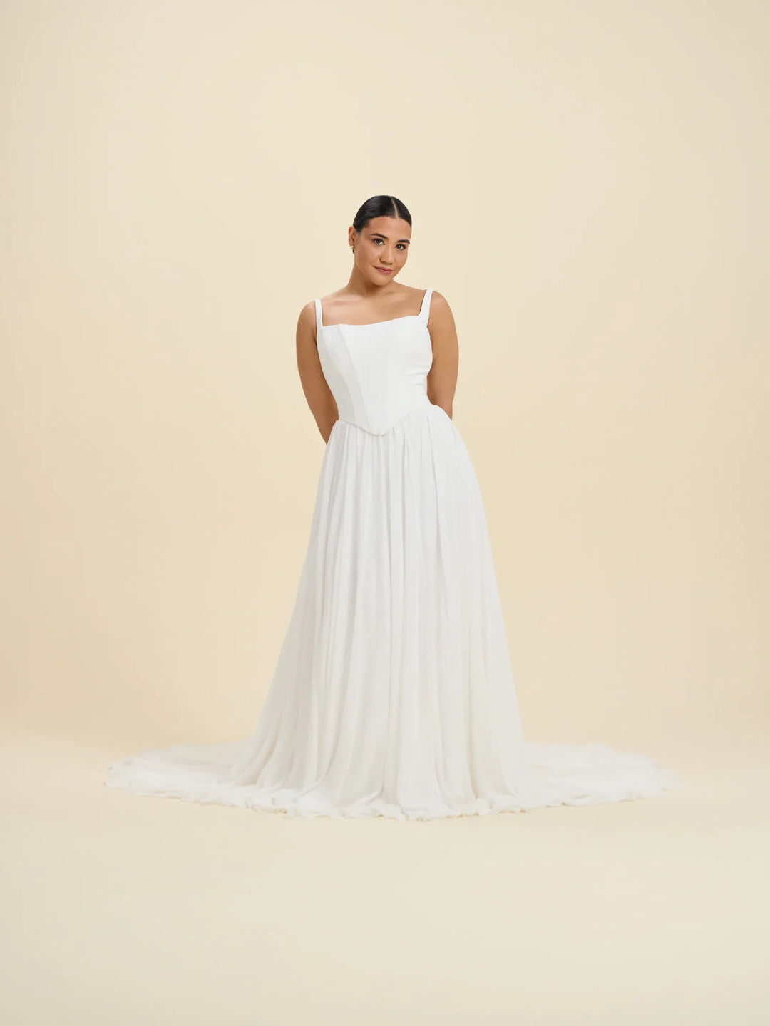 A woman modeling a white sleeveless A-line gown with a fitted bodice, square neckline, thin straps, and a long flowing skirt with a slight train, photographed against a plain warm beige studio background.