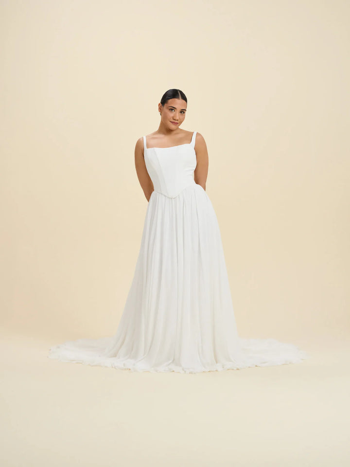 A woman modeling a white sleeveless A-line gown with a fitted bodice, square neckline, thin straps, and a long flowing skirt with a slight train, photographed against a plain warm beige studio background.