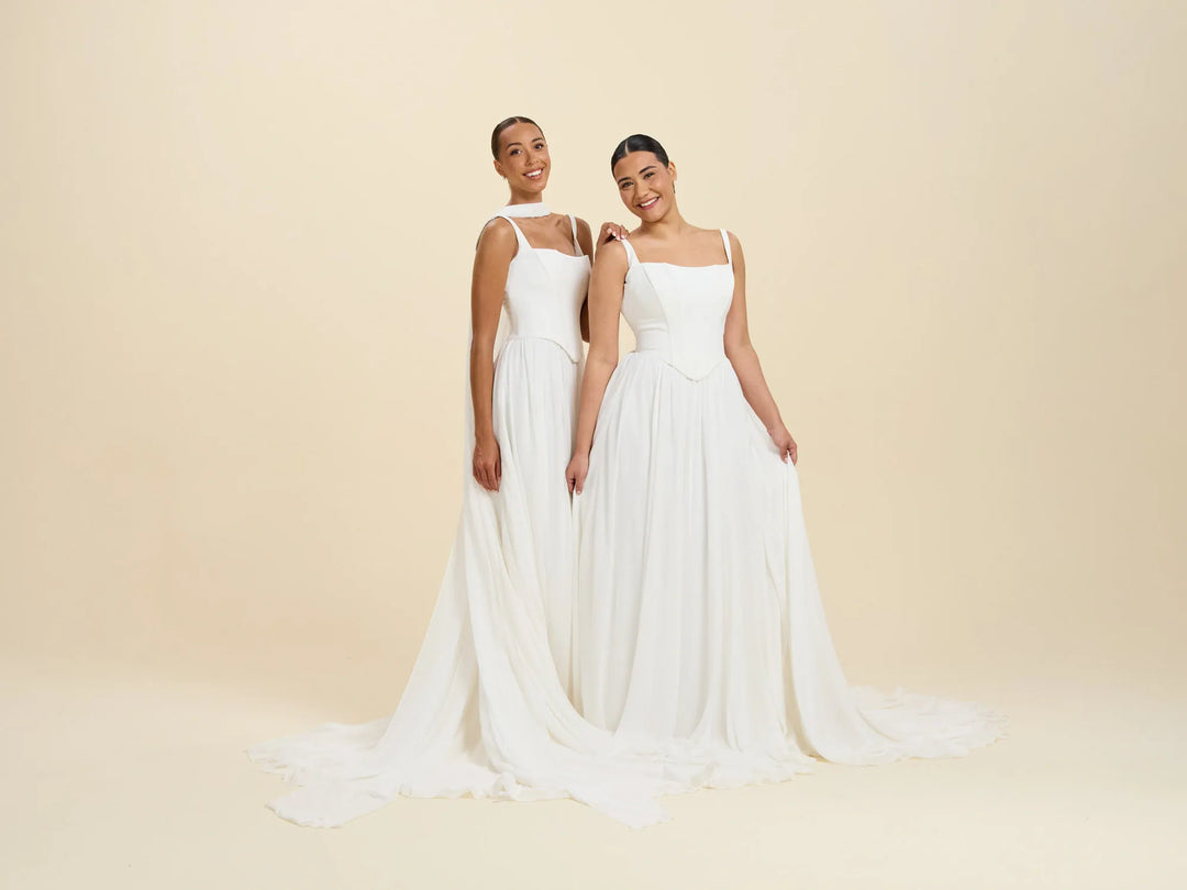 Two women modeling floor-length white wedding gowns with thin straps and flowing chiffon trains against a warm beige studio backdrop.
