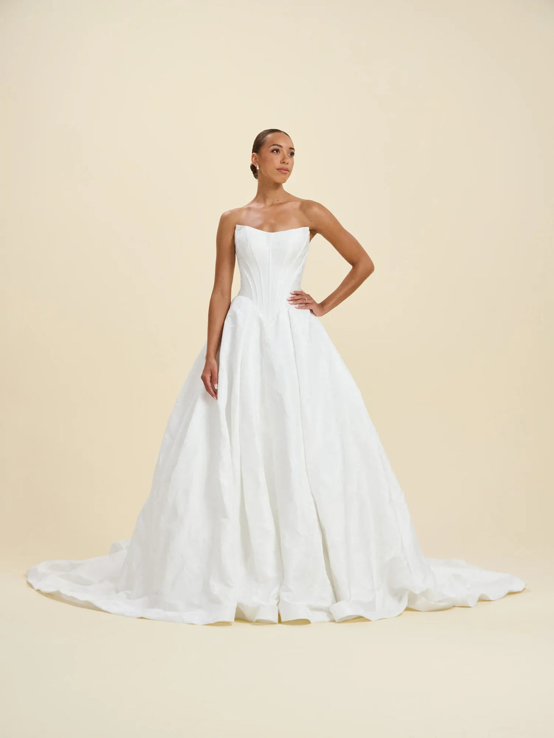 A woman modeling a strapless white wedding gown with a fitted bodice and full flowing skirt and train, posed against a plain warm beige studio background.