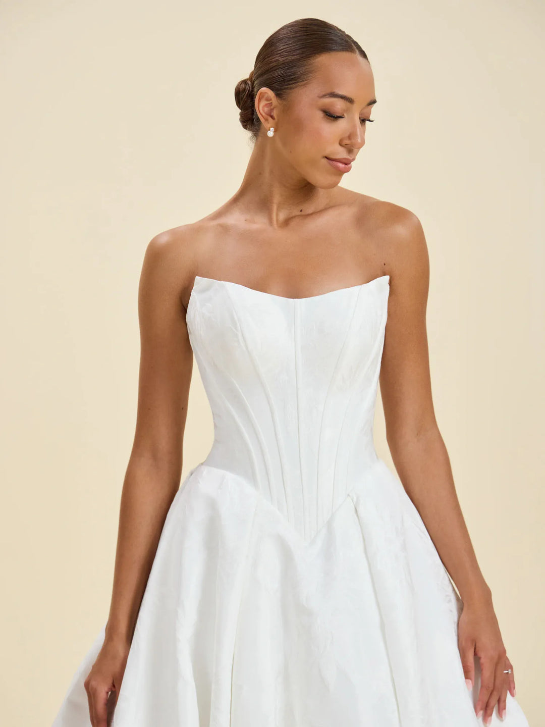 A woman modeling a strapless white wedding gown with a structured corset-style bodice and full skirt against a plain beige studio background.