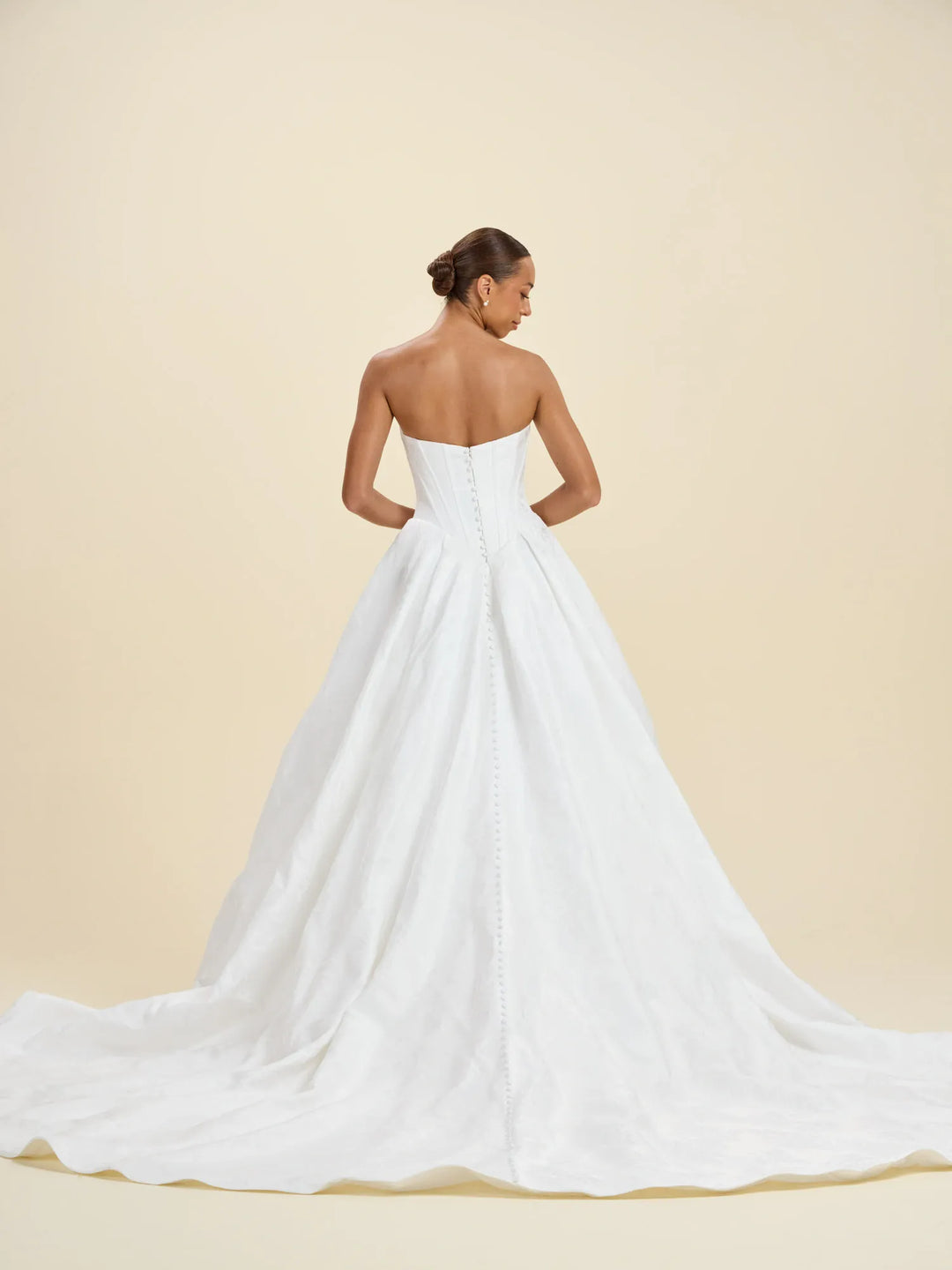 A woman modeling a strapless white wedding gown from the back, showing a structured corset-style back and a full ball gown with a long train, against a warm beige studio background.