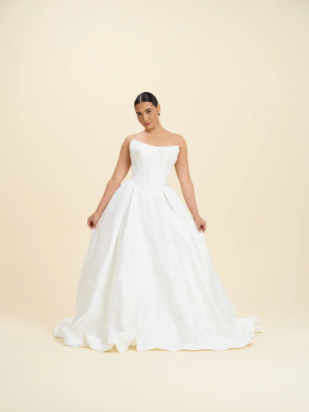 A woman modeling a strapless white ball gown wedding dress with a structured bodice and full skirt, standing against a plain beige studio background.