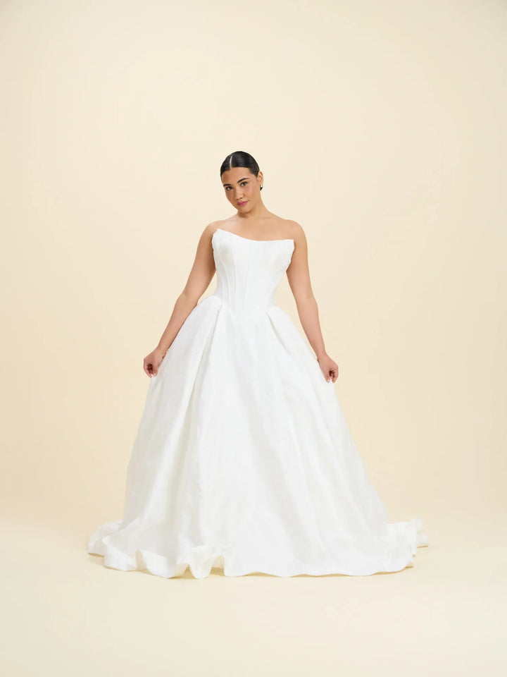 A woman modeling a strapless white ball gown wedding dress with a structured bodice and full skirt, standing against a plain beige studio background.