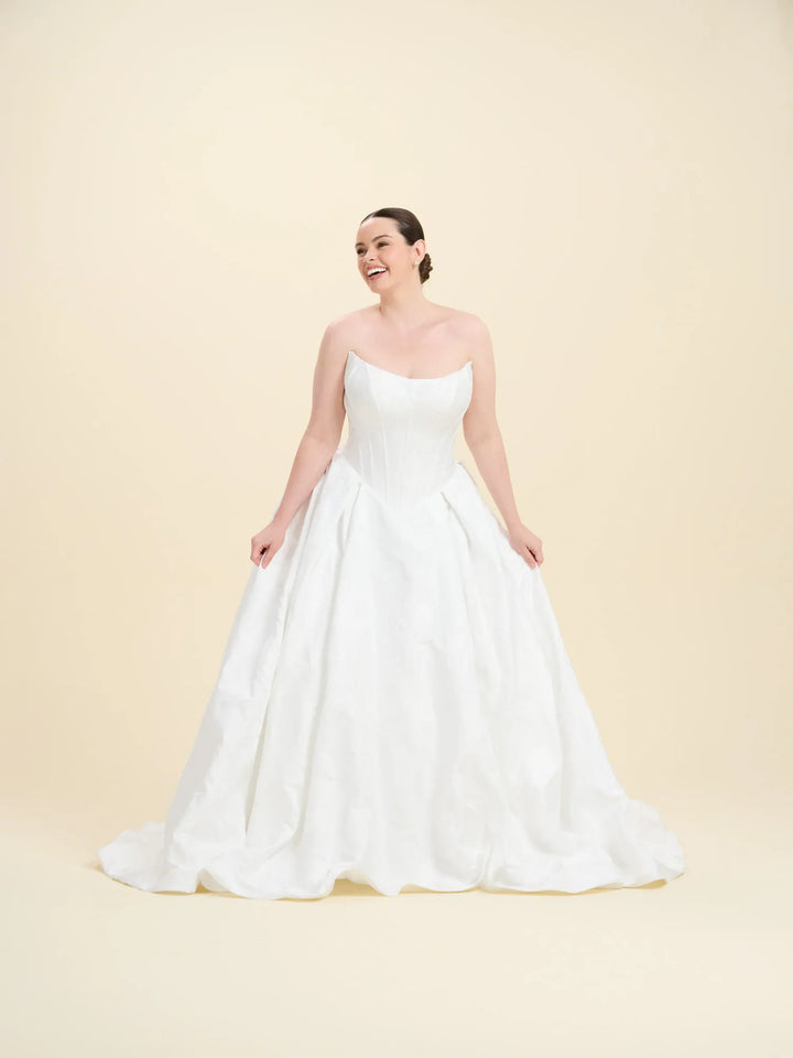 A woman modeling a strapless white ball gown wedding dress with a structured bodice and full skirt, standing against a solid light beige studio background.