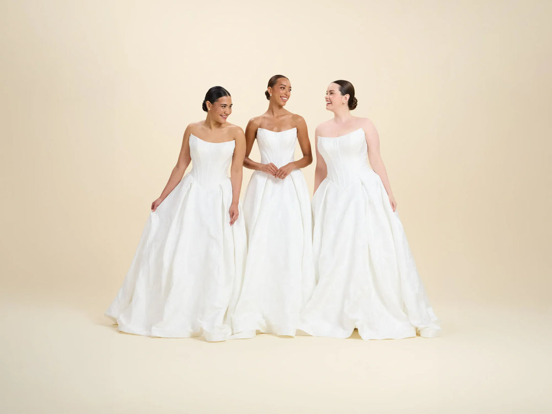 Three women wearing strapless white ballgown wedding dresses standing side by side against a warm beige studio background.