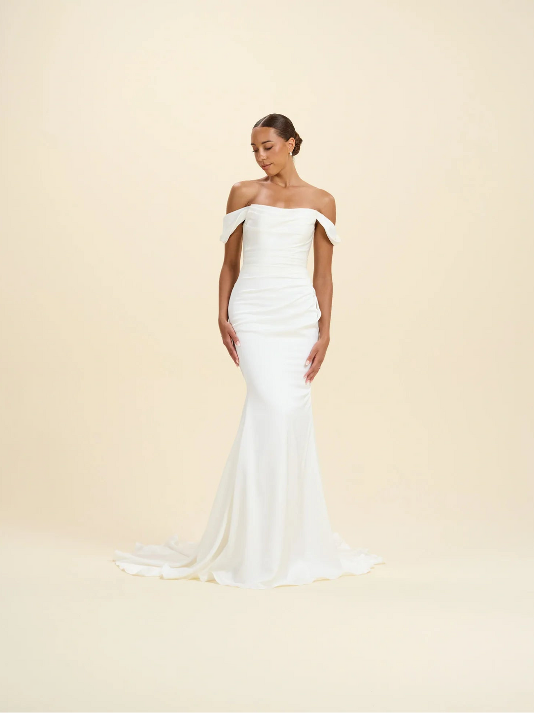 A woman modeling an off-the-shoulder white mermaid-style wedding gown with a fitted ruched bodice and a long flowing train, photographed against a plain warm cream studio background.