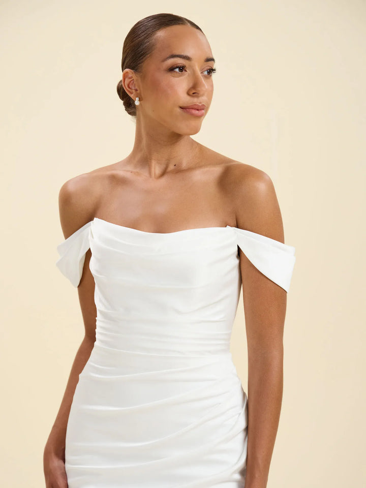 A woman wearing a white off-the-shoulder ruched dress, photographed against a solid light beige studio background.