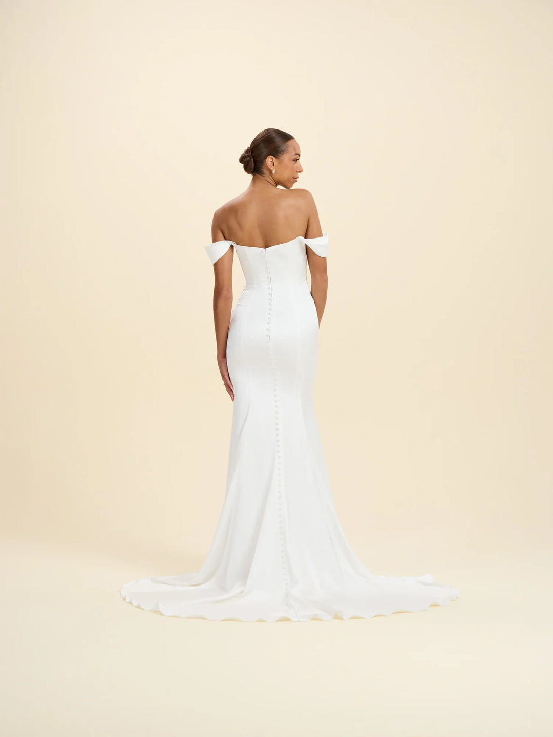 A woman modeling an off-the-shoulder white wedding gown from the back, standing against a solid warm cream studio background.