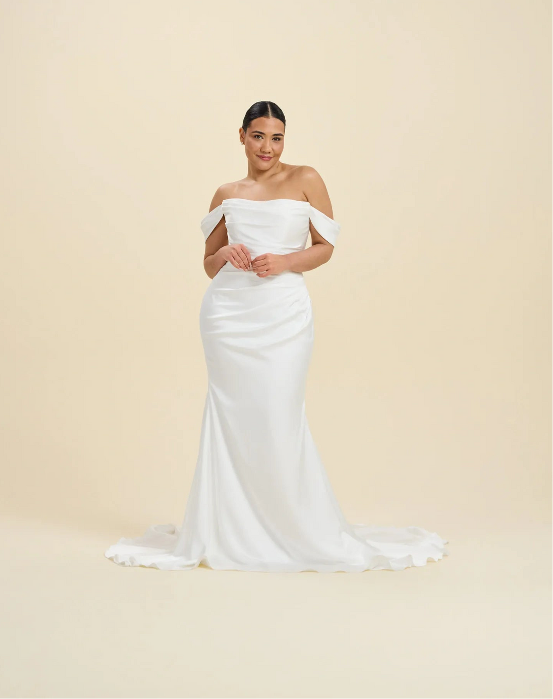 A woman modeling an off-the-shoulder white wedding gown with a fitted silhouette and a short train, standing against a plain beige studio background.