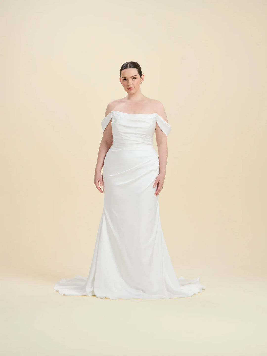 A woman modeling an off-the-shoulder white wedding gown with a fitted bodice, draped skirt, and slight train against a plain beige studio background.
