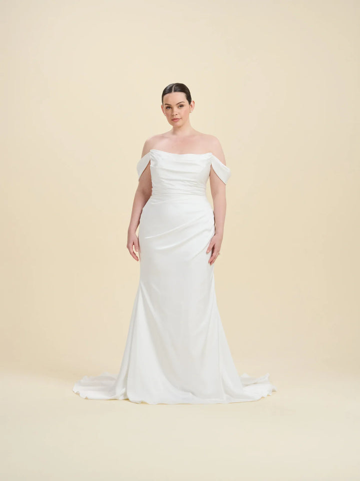 A woman modeling an off-the-shoulder white wedding gown with a fitted bodice, draped skirt, and slight train against a plain beige studio background.