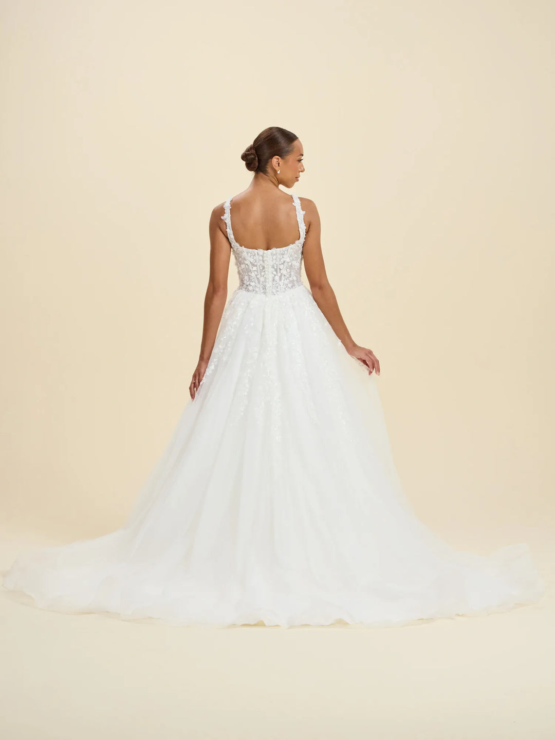 A woman modeling a white wedding gown from the back, showing a beaded lace bodice and a full tulle skirt, against a solid beige studio background.
