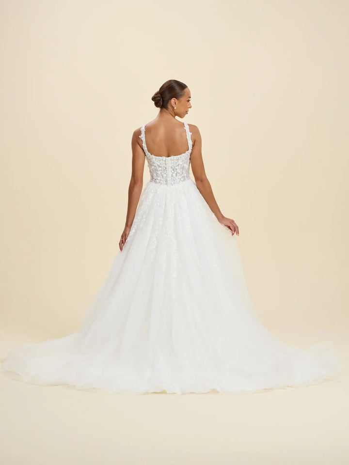 A woman modeling a white wedding gown from the back, showing a beaded lace bodice and a full tulle skirt, against a solid beige studio background.