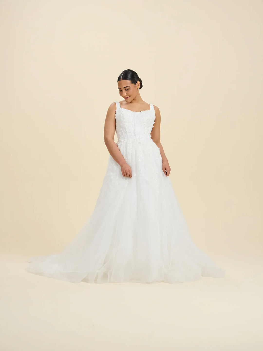A woman modeling a white sleeveless wedding gown with a fitted bodice, square neckline, and a full layered tulle skirt against a plain warm beige studio background.